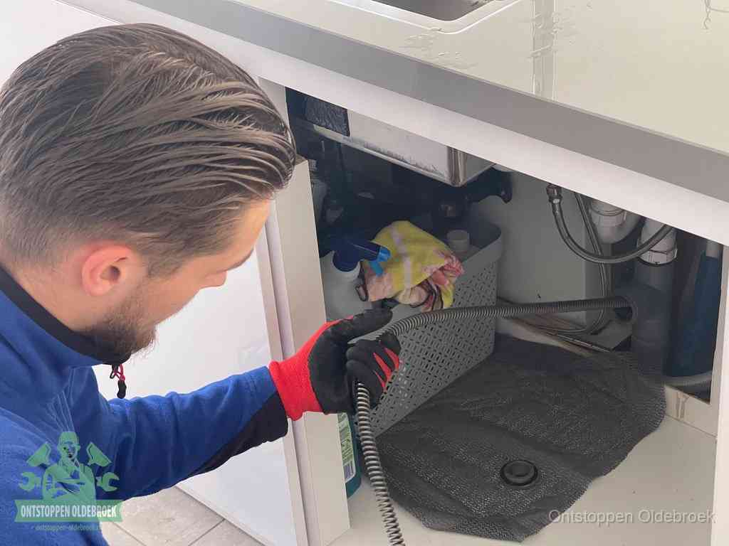 Een monteur voert een flexibele ontstoppingsveer in onder het keukenkastje, langs de sifon, om de gootsteen te ontstoppen in Oldebroek