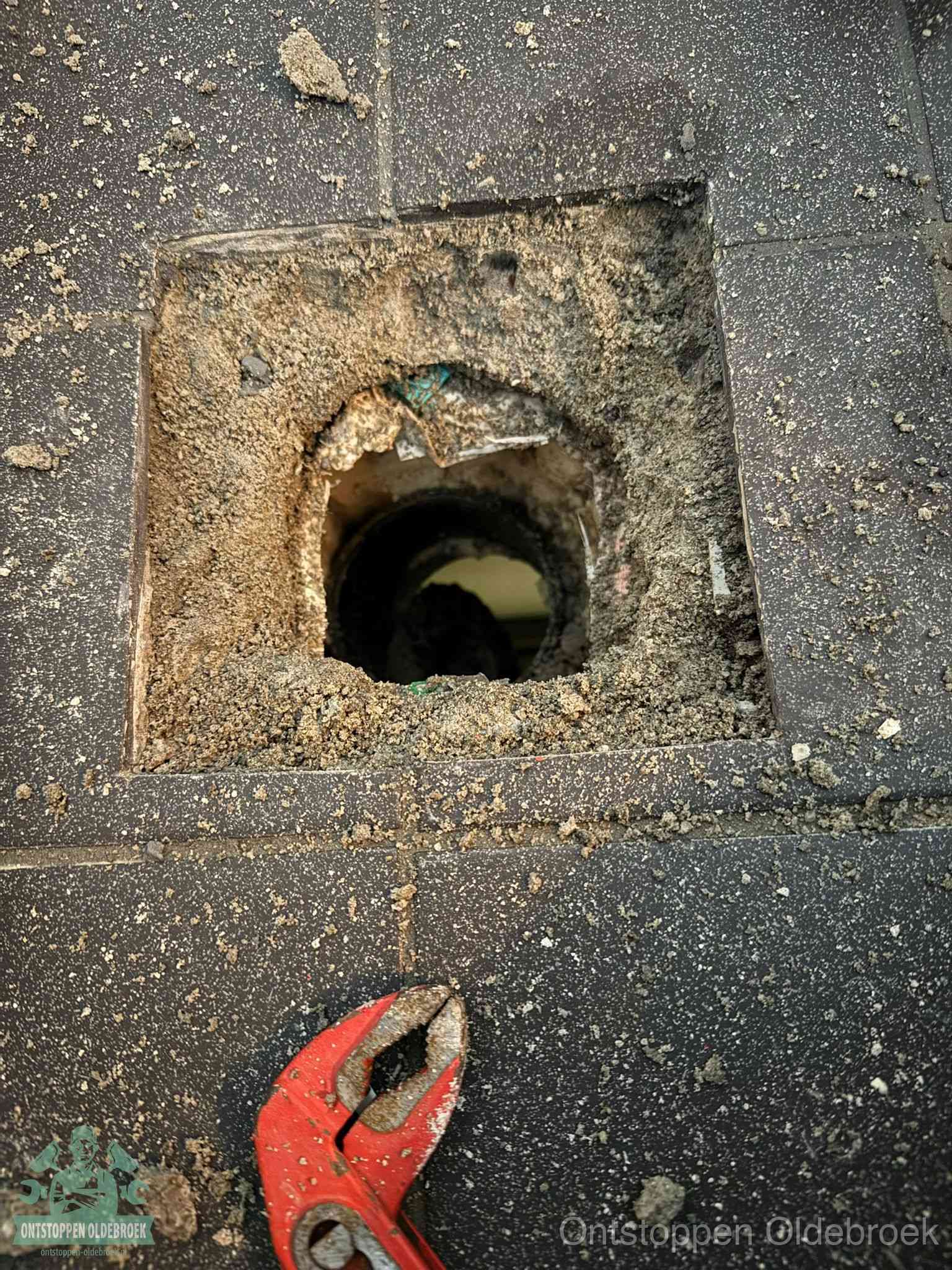 Een geopend rioolputje in een betontegelvloer met losgemaakt rooster en een zichtbare ronde afvoerbuis, het achterhalen waar de afvoerverstopping zit in Oldebroek