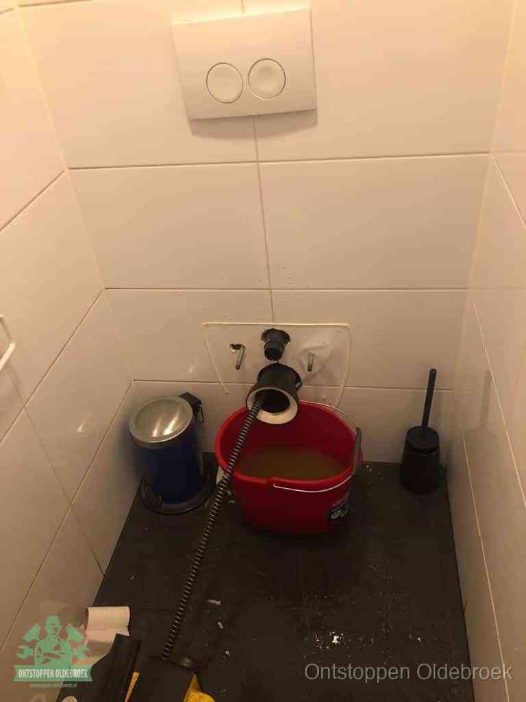 Een ontstopte wand-wc met verwijderde toiletpot waar een ontstoppingsveer in de wanduitgang verdwijnt, het achterhalen waar de verstopping zit in Oldebroek