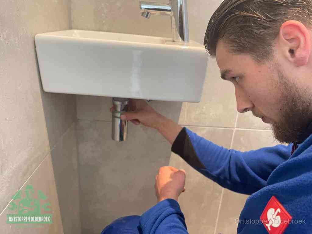 Een monteur draait met de hand de zwanenhalssifon onder een fonteinwasbak in de toiletruimte los om de bron van rioollucht in Oldebroek te vinden