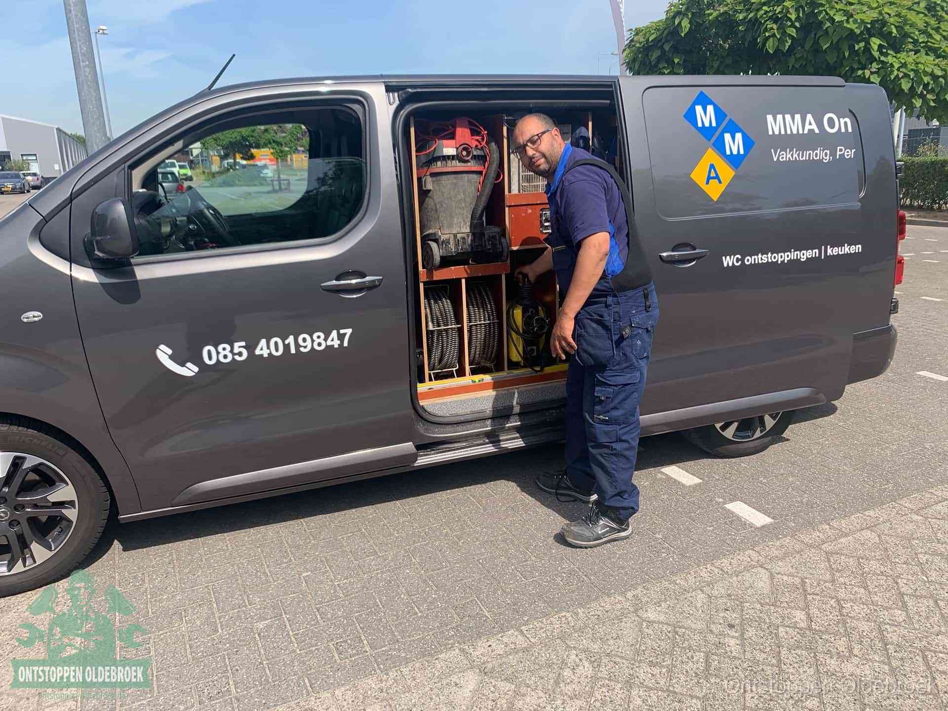 Een monteur bij de open schuifdeur van zijn bedrijfsbus op een parkeerplaats, klaar om de oorzaak van terugkerende afvoerverstoppingen aan te pakken in Oldebroek