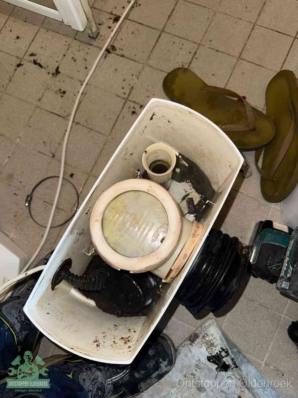 Een gedemonteerde toiletpot op de vloer met ontstoppingsveer en gereedschap ernaast, het verwijderen van een vastzittend voorwerp uit de wc in Oldebroek